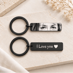 Two black rectangular keychains with round split rings, styled on a light beige stone surface. The top keychain has a black-and-white photo print showing a woman's eyes, and the bottom keychain has the white text 'I Love you' with a small heart.