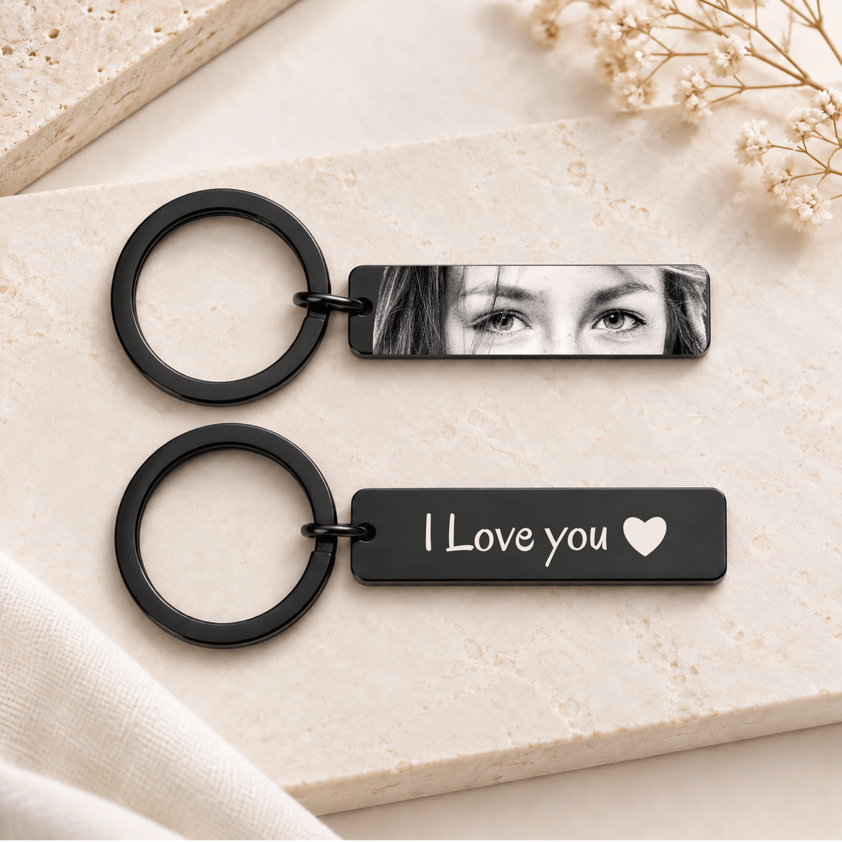 Two black rectangular keychains with round split rings, styled on a light beige stone surface. The top keychain has a black-and-white photo print showing a woman's eyes, and the bottom keychain has the white text 'I Love you' with a small heart.