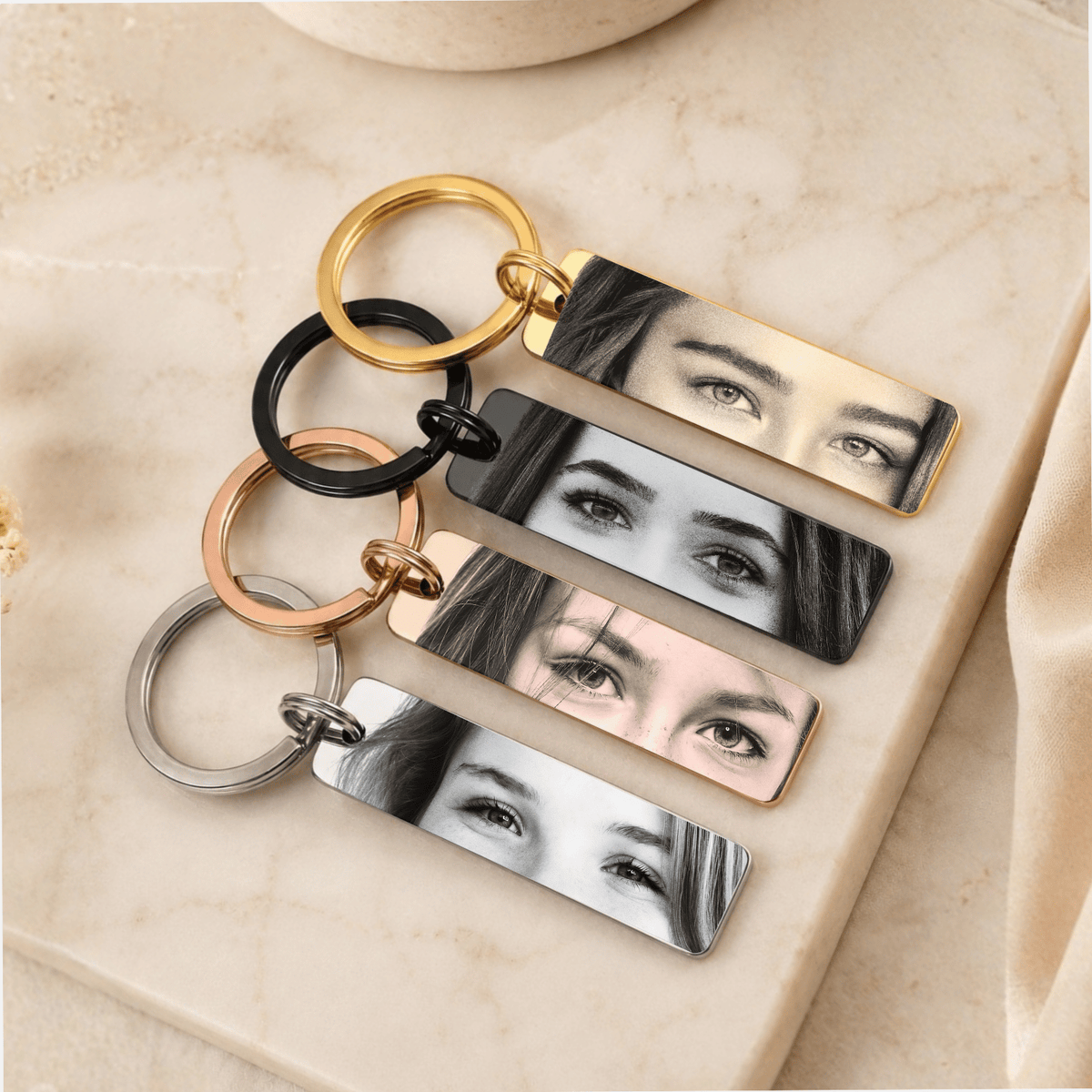 Four rectangular metal keychains with custom photo prints of a woman's eyes, shown in gold, black, rose gold, and silver finishes on a beige marble surface.