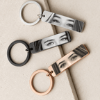 Three rectangular keychains with a glossy black-and-white portrait of a woman's eyes are arranged diagonally on a light beige textured surface. The keychains come in silver, black, and rose gold metal finishes.