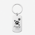 A personalized silver dog tag keychain with a circular split ring, featuring black paw prints, the name 'Poppy' in cursive, and the date '07.04.
