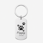 A personalized silver dog tag keychain with a circular split ring, featuring black paw prints, the name 'Poppy' in cursive, and the date '07.04.