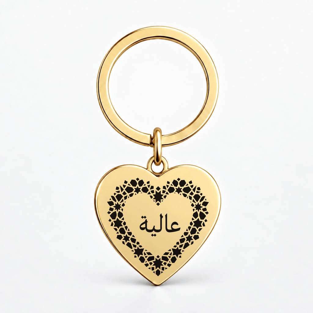 A gold heart-shaped keychain with a black floral border and Arabic script on a clean white background.