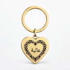 A gold heart-shaped keychain with a black floral border and Arabic script on a clean white background.