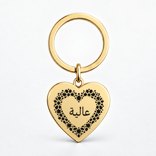 A gold heart-shaped keychain with a black floral border and Arabic script on a clean white background.