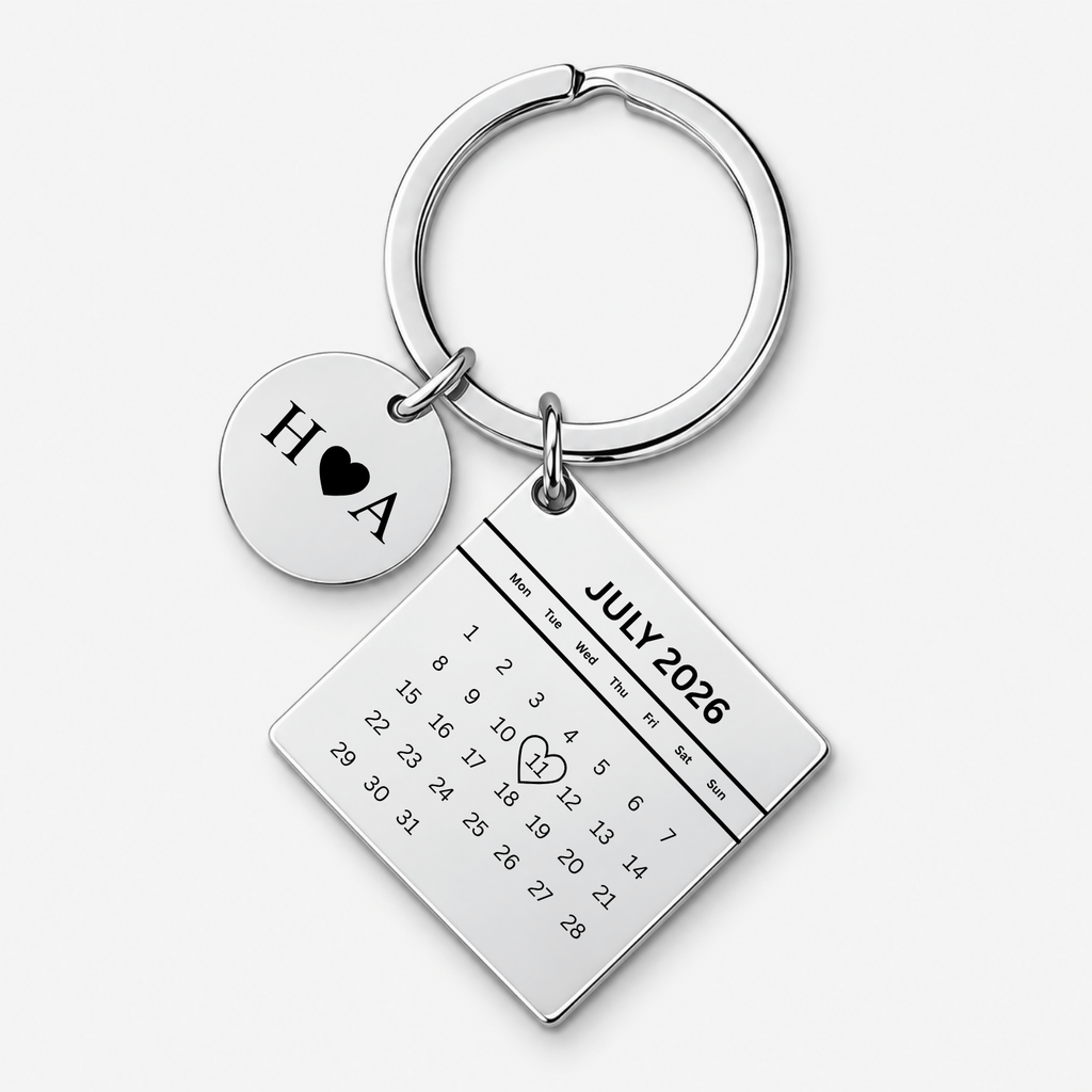 A silver-toned metal keychain featuring a round disc charm engraved with 'H A' and a black heart, alongside a rectangular calendar charm displaying 'JULY 2026' with a small heart drawn over the number 14. The keychain is photographed on a clean, light gray background.