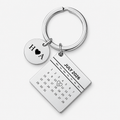 A silver-toned metal keychain featuring a round disc charm engraved with 'H A' and a black heart, alongside a rectangular calendar charm displaying 'JULY 2026' with a small heart drawn over the number 14. The keychain is photographed on a clean, light gray background.