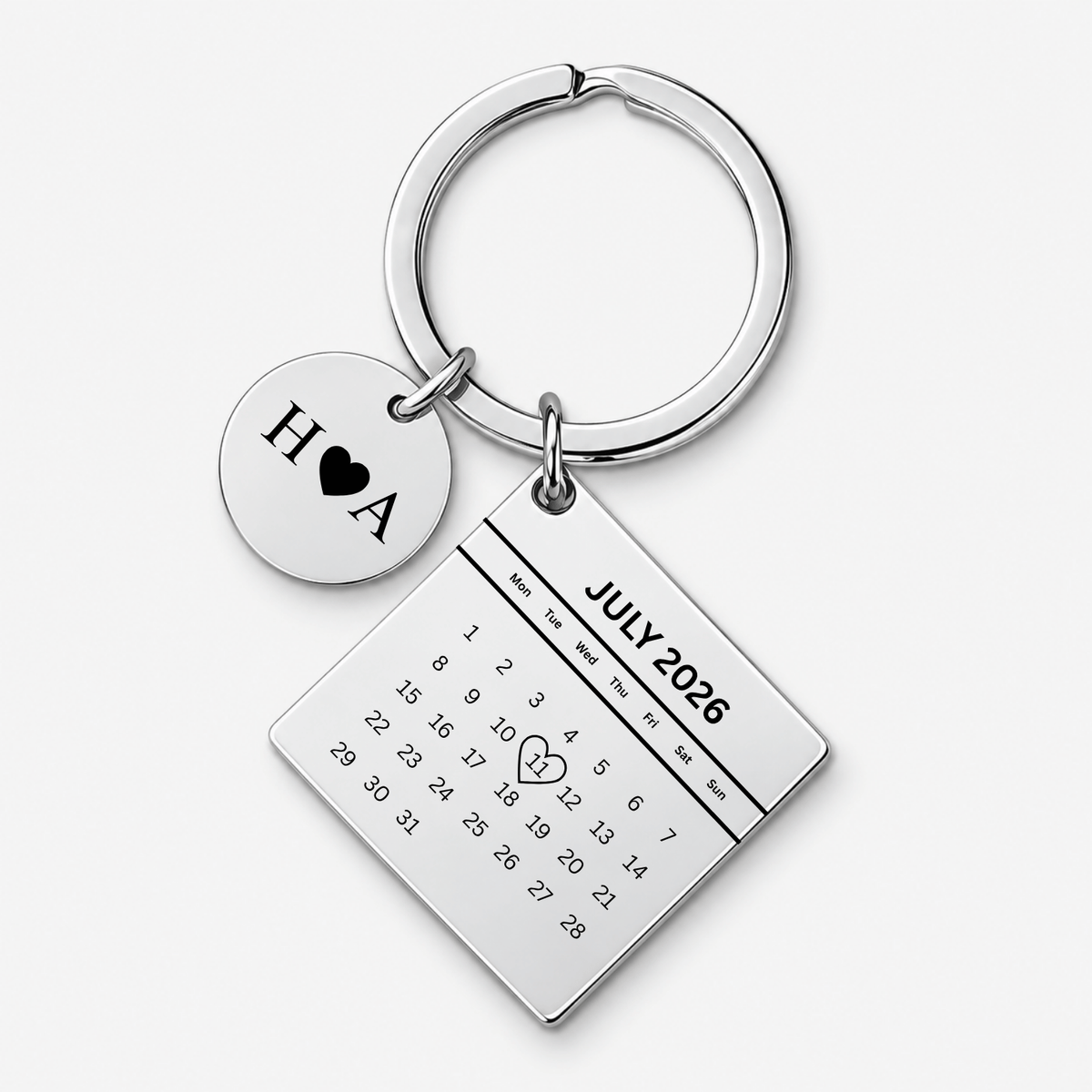 A silver-toned metal keychain featuring a round disc charm engraved with 'H A' and a black heart, alongside a rectangular calendar charm displaying 'JULY 2026' with a small heart drawn over the number 14. The keychain is photographed on a clean, light gray background.