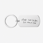 A silver metal keychain with a rectangular tag engraved with the handwritten text 'always here for you, from Mum's Dad' on a light gray background.